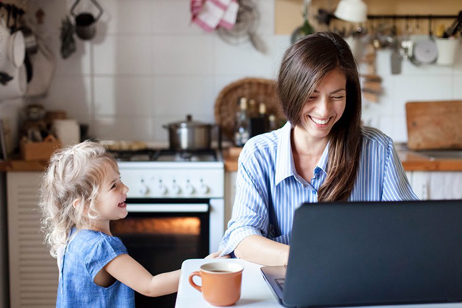 Blog - Happy Mother and Daughter Smiling While Mother Blogs on Her Laptop in the Kitchen
